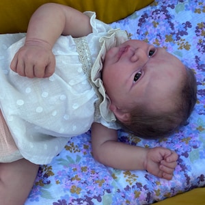 Puede incluir: Una mu&ntilde;eca beb&eacute; realista con un vestido blanco con un patr&oacute;n de puntos y adornos de encaje. La mu&ntilde;eca est&aacute; acostada sobre una manta con estampado floral con flores moradas y azules. La piel de la mu&ntilde;eca tiene un tono realista.
