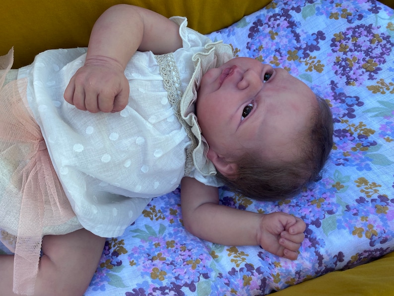 Puede incluir: Una mu&ntilde;eca beb&eacute; realista con un vestido blanco con un patr&oacute;n de puntos y adornos de encaje. La mu&ntilde;eca est&aacute; acostada sobre una manta con estampado floral con flores moradas y azules. La piel de la mu&ntilde;eca tiene un tono realista.