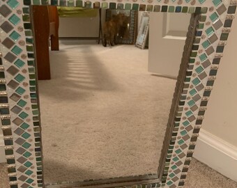 May include: Square mirror with a mosaic tile frame. The frame has a pattern of teal, grey, and clear tiles, with small decorative accents. The mirror is leaning against a wall.