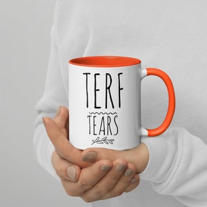 May include: White ceramic mug with an orange rim and handle. The mug has black text that reads "TERF TEARS" with a small floral design below the text.