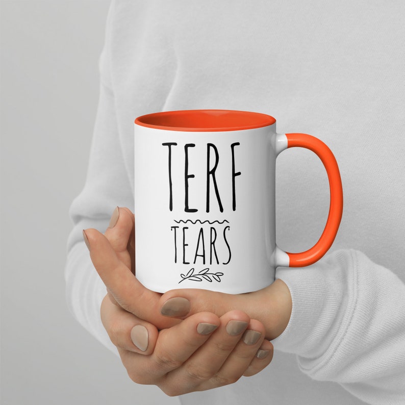 May include: White ceramic mug with an orange rim and handle. The mug has black text that reads "TERF TEARS" with a small floral design below the text.