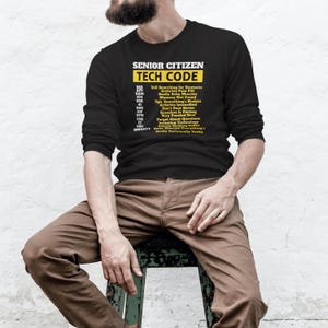 May include: Black long-sleeve shirt with the text "SENIOR CITIZEN TECH CODE" in yellow and white. The shirt lists humorous tech-related acronyms and phrases. The person is sitting on a wooden stool.