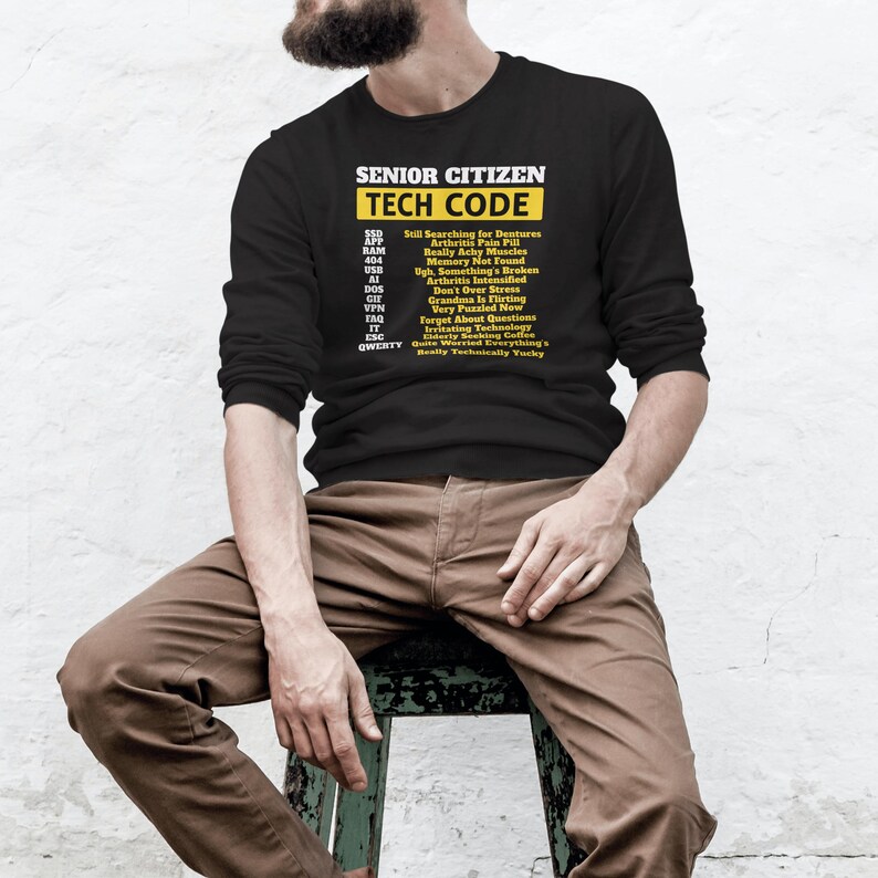 May include: Black long-sleeve shirt with the text "SENIOR CITIZEN TECH CODE" in yellow and white. The shirt lists humorous tech-related acronyms and phrases. The person is sitting on a wooden stool.