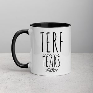 May include: White ceramic mug with a black rim and handle. The mug has black text that reads "TERF TEARS" with a small floral design below the text.