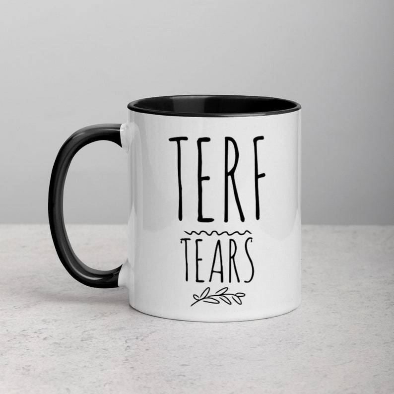 May include: White ceramic mug with a black rim and handle. The mug has black text that reads "TERF TEARS" with a small floral design below the text.