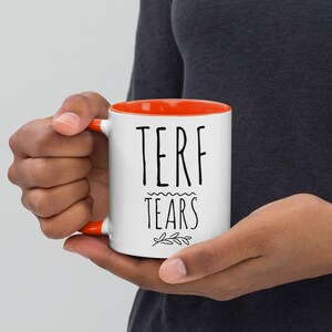 May include: White ceramic mug with an orange rim. The mug has black text that reads "TERF TEARS" with a small floral design underneath.