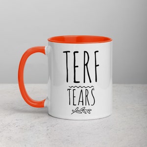 May include: White ceramic mug with an orange rim. The mug has black text that reads "TERF TEARS" with a small branch design below the text.