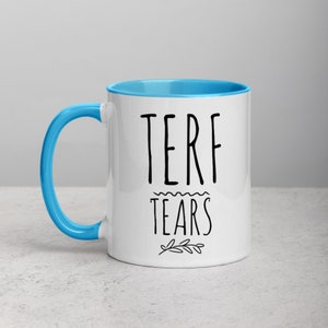 May include: White ceramic mug with a blue rim and handle. The mug has black text that reads "TERF TEARS" with a small floral design underneath.