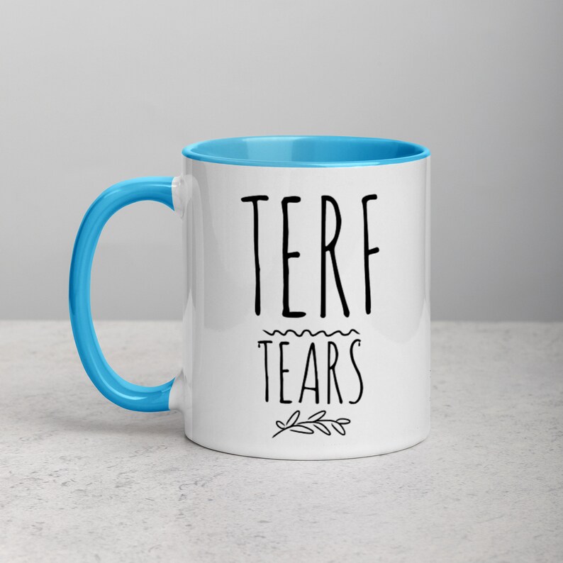 May include: White ceramic mug with a blue rim and handle. The mug has black text that reads "TERF TEARS" with a small floral design underneath.