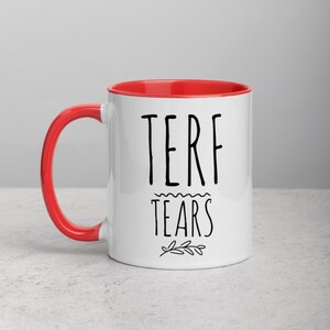 May include: White ceramic mug with a red rim. The mug has black text that reads "TERF TEARS" with a small floral design below the text.