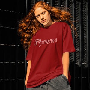 Puede incluir: Una camiseta roja con un contorno blanco de la palabra "PYTRON" en la parte delantera.