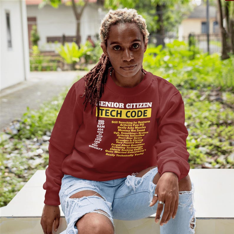 May include: A maroon sweatshirt with the text "SENIOR CITIZEN TECH CODE" in white and yellow. Below the title are tech-related acronyms and phrases like "Arthritis Pain Pill" and "Memory Not Found". The person is wearing ripped blue jeans.