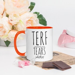 May include: White ceramic mug with an orange rim. The mug has black text that reads "TERF TEARS" with a small branch design below the text.