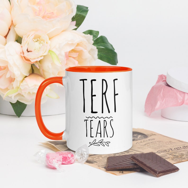 May include: White ceramic mug with an orange rim. The mug has black text that reads "TERF TEARS" with a small branch design below the text.
