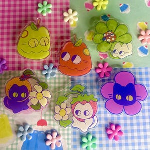 Puede incluir: Una colección de pegatinas coloridas y caprichosas con diseños de frutas y flores con caras de gato. Las pegatinas incluyen una manzana, una naranja, una flor con una mariquita y una fresa. Adornos decorativos de flores y cupcakes están dispersos.