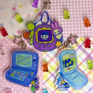 Könnte beinhalten: Acryl-Schlüsselanhänger mit Cartoon-Designs eines Handheld-Gaming-Geräts, eines Klapphandys und eines virtuellen Haustiers im Tamagotchi-Stil. Jeder Schlüsselanhänger hat einen sternförmigen Verschluss und ist von bunten Gummibärchen umgeben.