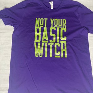Puede incluir: Camiseta morada con el texto "NOT YOUR BASIC WITCH" en verde.