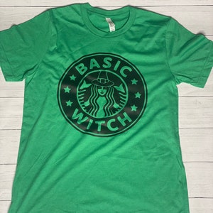 May include: Green short-sleeved t-shirt with a black graphic of a witch wearing a hat and a coffee cup with the text "BASIC WITCH" around it.