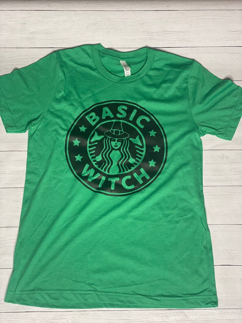 May include: Green short-sleeved t-shirt with a black graphic of a witch wearing a hat and a coffee cup with the text "BASIC WITCH" around it.