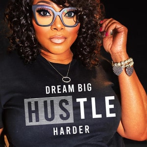 Może przedstawiać: Czarny T-shirt z białym tekstem, który brzmi "Dream Big Hustle Harder". Tekst jest pogrubiony, bezszeryfowy i otoczony białą, paskowaną ramką.