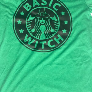 May include: Green t-shirt with a black and white graphic of a witch wearing a hat and the text "BASIC WITCH" in a circle.