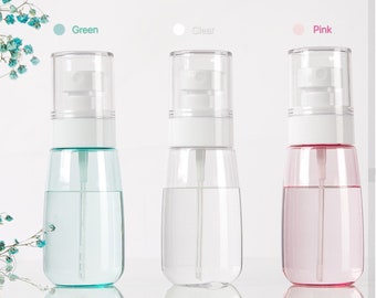 May include: Three empty spray bottles with clear plastic bodies and white tops. The bottles are in three colors: green, clear, and pink. The bottles are on a white surface.