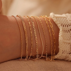 Peut inclure: Un gros plan d'un poignet portant une pile de bracelets en chaîne dorés. Les bracelets sont de différentes largeurs et de différents styles, créant un look superposé.