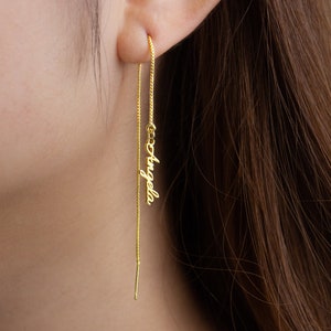 Pode incluir: Um brinco de fio dourado personalizado com o nome "Angela" em uma escrita delicada. O brinco é usado na orelha, com uma corrente longa e uma pequena barra na extremidade. O fundo está desfocado, focando na joia.