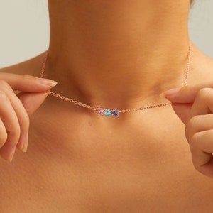 Może przedstawiać: Delikatny złoty naszyjnik z trzema małymi kamieniami szlachetnymi: różowym, niebieskim i fioletowym. Naszyjnik nosi osoba, ale skupienie jest na samym naszyjniku.