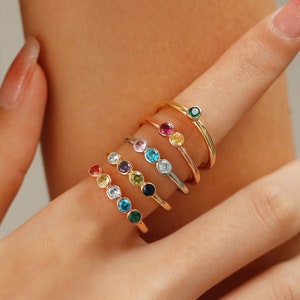 Op de afbeelding: Een set van zeven gouden ringen met gekleurde edelstenen. Elke ring heeft een enkele ronde edelsteen in een andere kleur, waaronder rood, roze, geel, groen, blauw, turquoise en oranje.
