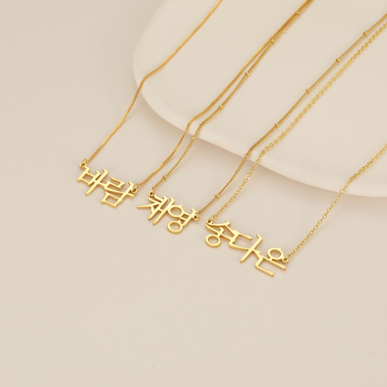 May include: Three gold necklaces with Korean words hanging from them. The words are "바람", "채영", and "승다은".
