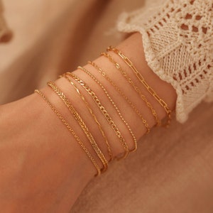 Peut inclure: Un ensemble de neuf bracelets en chaîne dorés superposés au poignet d'une personne. Les bracelets sont de styles et d'épaisseurs différents.