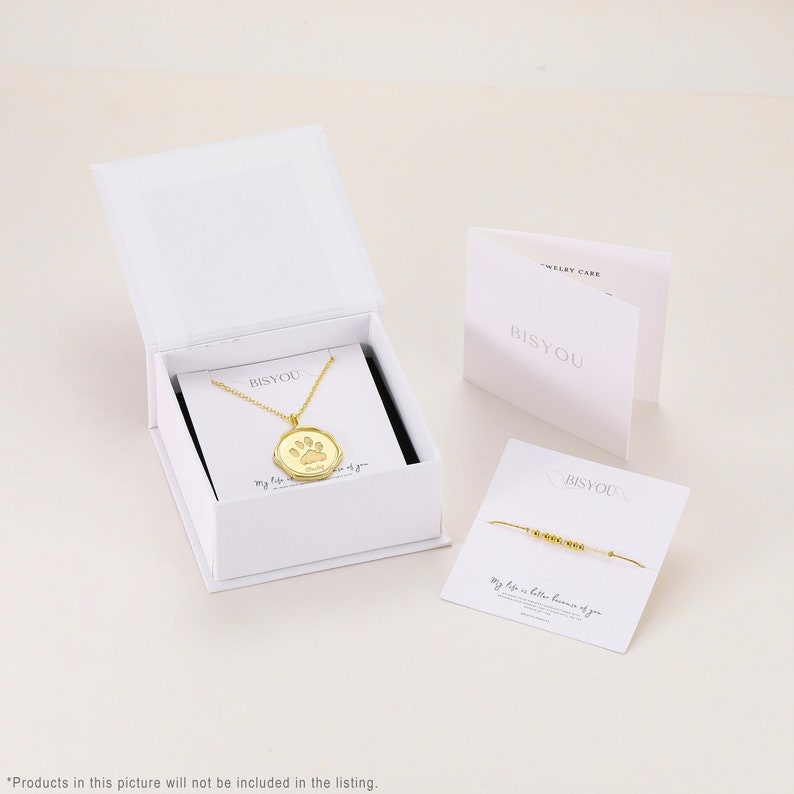 May include: A white box with a gold necklace and a paw print pendant. The necklace is in a white box with the word "BISYOU" printed on it. The box is sitting on a white surface. The necklace is accompanied by a bracelet with the words "My life is better because of you" printed on it.