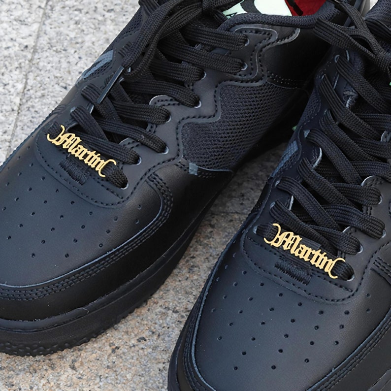 May include: Close-up of two black sneakers with black laces. The laces have gold metal charms with the word "Martin" written in a script font.