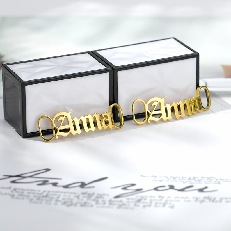 May include: Two gold-colored nameplates with the name "Anna" spelled in a gothic font. The nameplates are on a white surface.