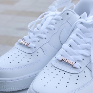 May include: A pair of white leather sneakers with white laces and rose gold metal lace charms that say "Mike".