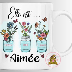 Peut inclure: Mug en céramique blanche avec un motif floral et le texte "Elle est... Aimée" en noir. Le motif floral présente trois bocaux en verre remplis de fleurs colorées et de papillons.
