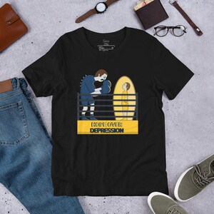 May include: Black t-shirt with a graphic design featuring a boxer facing a child inside a boxing ring. The text "HOPE OVER DEPRESSION" is displayed on a yellow banner. The shirt has a crew neck and short sleeves.