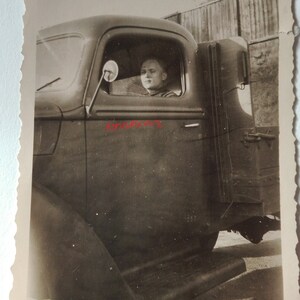 May include: A black and white photograph of a person sitting in the driver's seat of a lorry. The person is looking straight ahead and has a serious expression on their face. The lorry is parked on a dirt road and there is a wooden fence in the background.