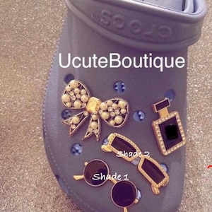 May include: A light purple clog shoe featuring the text UcuteBoutique and four decorative charms: a pearl-encrusted bow, a rectangular perfume bottle, and two pairs of sunglasses labeled Shade 1 and Shade 2, all with gold-tone metal accents.