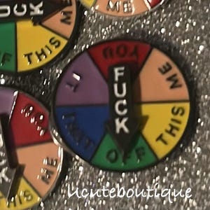 May include: A circular metal enamel pin featuring a spinning arrow pointing to segments colored red, peach, yellow, green, blue, and purple. Each segment contains black text, and the central arrow is black with the white text FUCK printed vertically.