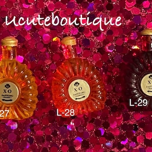 May include: Three miniature glass bottles with gold caps, labeled L-27, L-28, and L-29. Each bottle features a round white label with the text X.O FASTKING. The bottles are colored yellow, orange, and dark brown, with a ribbed, fan-like design.