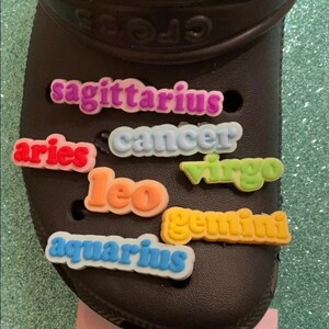 May include: A black pair of clogs with colorful zodiac sign charms attached. The charms spell out "sagittarius", "cancer", "aries", "leo", "virgo", "gemini", and "aquarius".