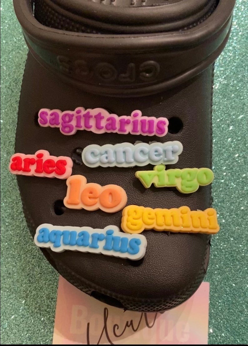 May include: A black pair of clogs with colorful zodiac sign charms attached. The charms spell out "sagittarius", "cancer", "aries", "leo", "virgo", "gemini", and "aquarius".