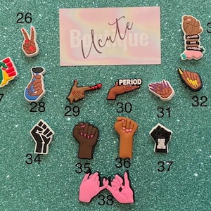 May include: A collection of shoe charms featuring various hand gestures, raised fists, and social justice symbols, each numbered from 26 to 38. The charms display diverse skin tones, colorful manicures, and text including PERIOD and BLACK LIVES MATTER.
