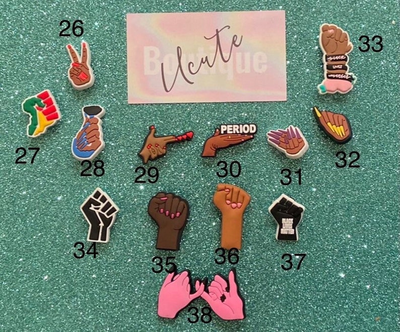 May include: A collection of shoe charms featuring various hand gestures, raised fists, and social justice symbols, each numbered from 26 to 38. The charms display diverse skin tones, colorful manicures, and text including PERIOD and BLACK LIVES MATTER.