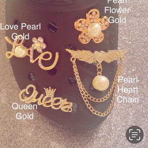 May include: A collection of four gold-toned decorative shoe charms featuring a script word Love with a pearl, a flower with a central pearl, a script word Queen with a crown, and a winged heart with a dangling pearl and gold chain.
