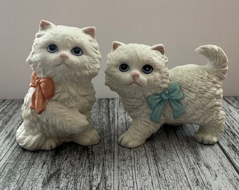 Puede incluir: Dos figuritas de gato de porcelana blanca con pelaje esponjoso. Un gato tiene un lazo de color melocotón y el otro tiene un lazo azul claro.