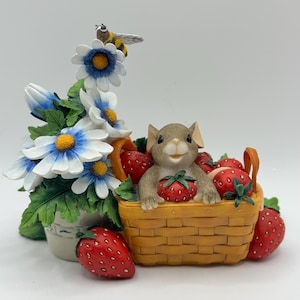 May include: A whimsical figurine featuring a smiling mouse in a woven basket filled with red strawberries. A bee rests on a cluster of white and blue daisies. The figurine is a charming decorative piece.