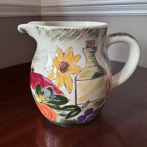 May include: A white ceramic pitcher with a handle, featuring a painted design of sunflowers, grapes, leaves, and a bottle. The pitcher has a rustic, farmhouse style.